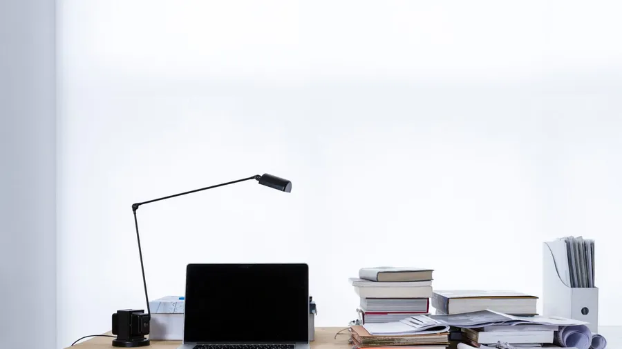 Small white desk with a laptop, a desk lamp and a stack of books against a white wall