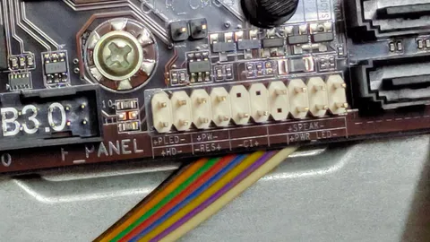 Close-up of a desktop motherboard's front panel header with a multicoloured ribbon cable attached, next to a clear CMOS jumper