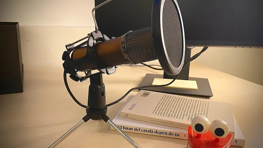 USB microphone with a pop filter on a small tripod stand, on a desk beside a monitor and a stack of books