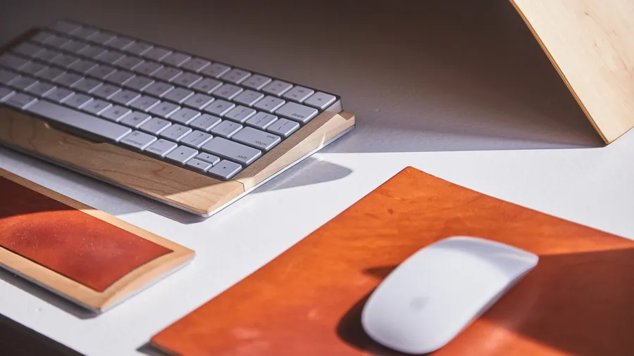 Wireless mouse on a leather desk pad next to a keyboard on a wooden stand